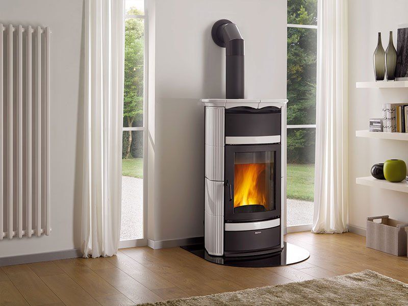Una moderna stufa a legna con un fuoco scoppiettante in un luminoso soggiorno accanto a grandi finestre e tende.