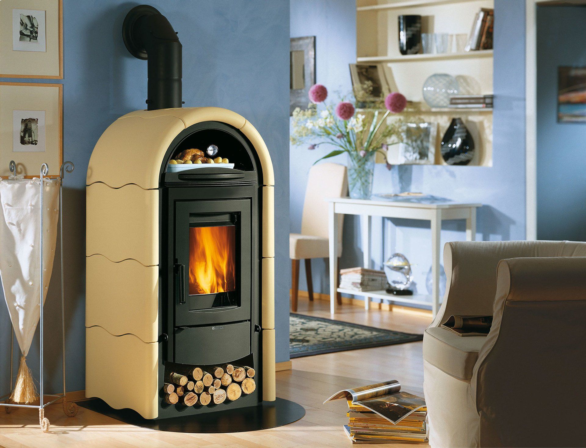 Soggiorno moderno con stufa a legna color crema e nera, camino acceso e ceppi di legno accatastati sotto.