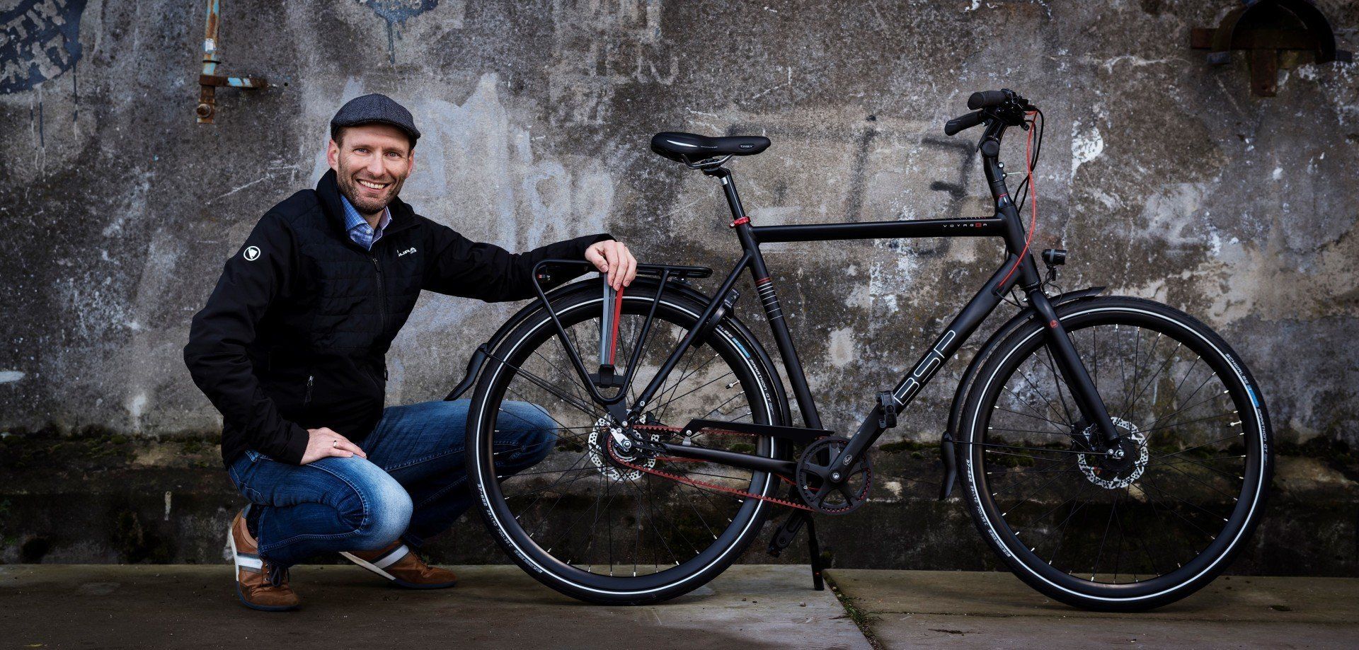 Een man knielt naast een zwarte fiets tegen een verweerde muur, glimlachend en met een hand op het achterwiel.