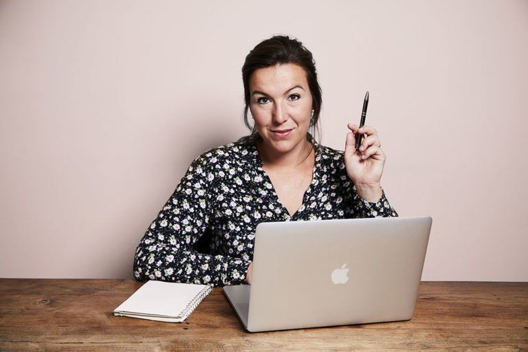 Vrouw zit achter een bureau met een laptop, glimlachend en met een pen naast een notitieboekje.