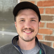 A man wearing a hat and a jacket is smiling in front of a brick wall.