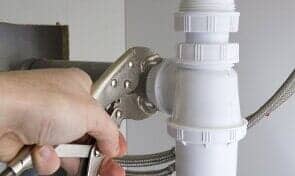 Hand using pliers to tighten a white plastic plumbing connection under a sink.