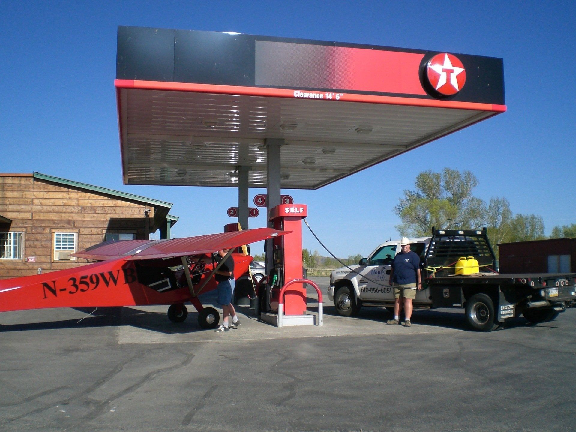 Red plane refueling at a Texaco gas station next to a flatbed truck; sunny day.
