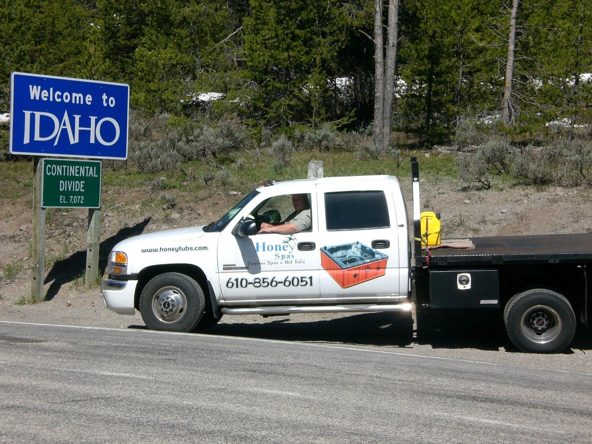 White truck with Idaho sign. Driver in cab, flatbed. Landscape with trees.