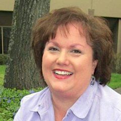 Woman with brown hair smiles outdoors in a light purple shirt, with a tree and building in background.