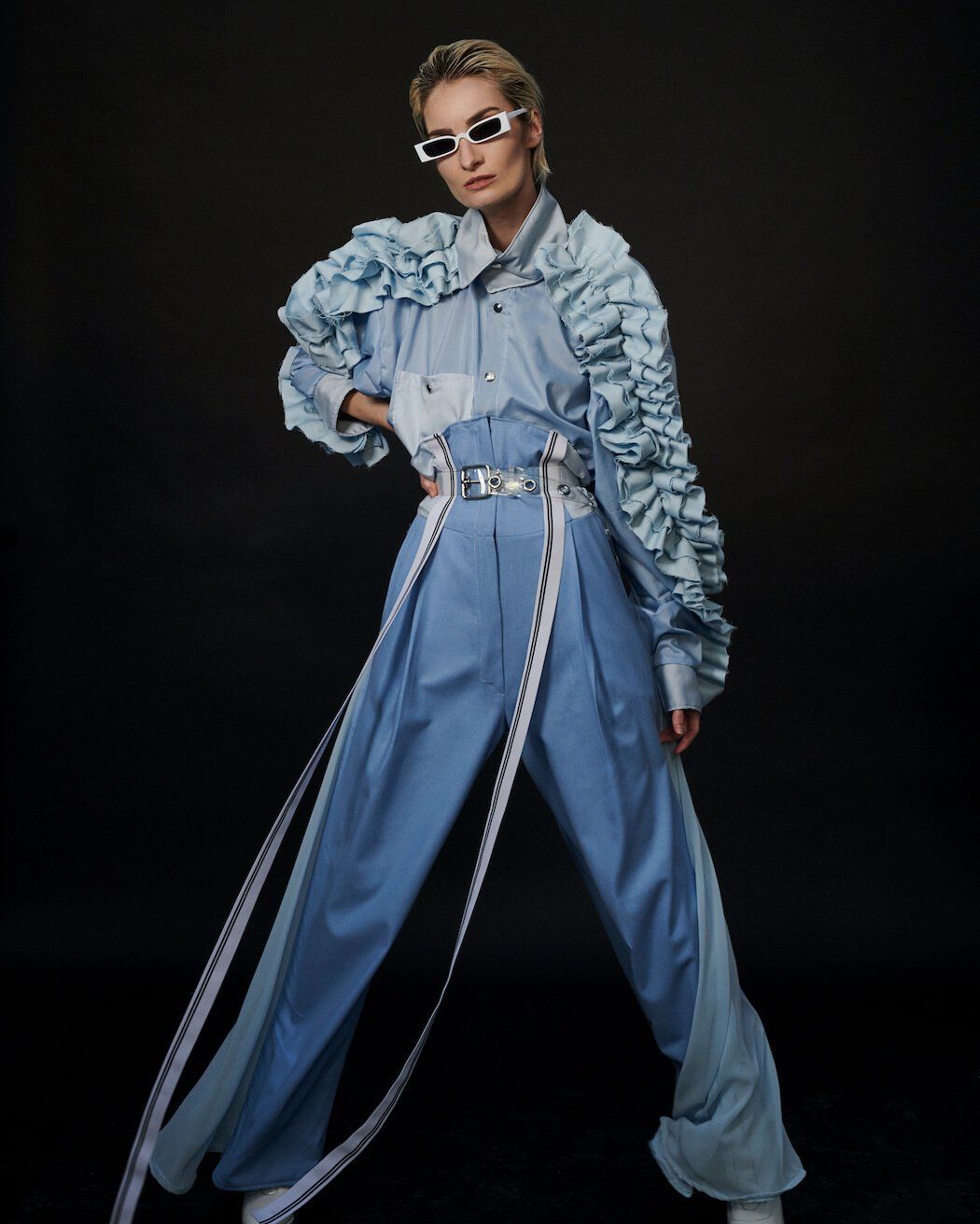 Frau in stylischem blauem Jeans-Outfit mit Rüschenärmeln und Sonnenbrille, posiert vor schwarzem Hintergrund.