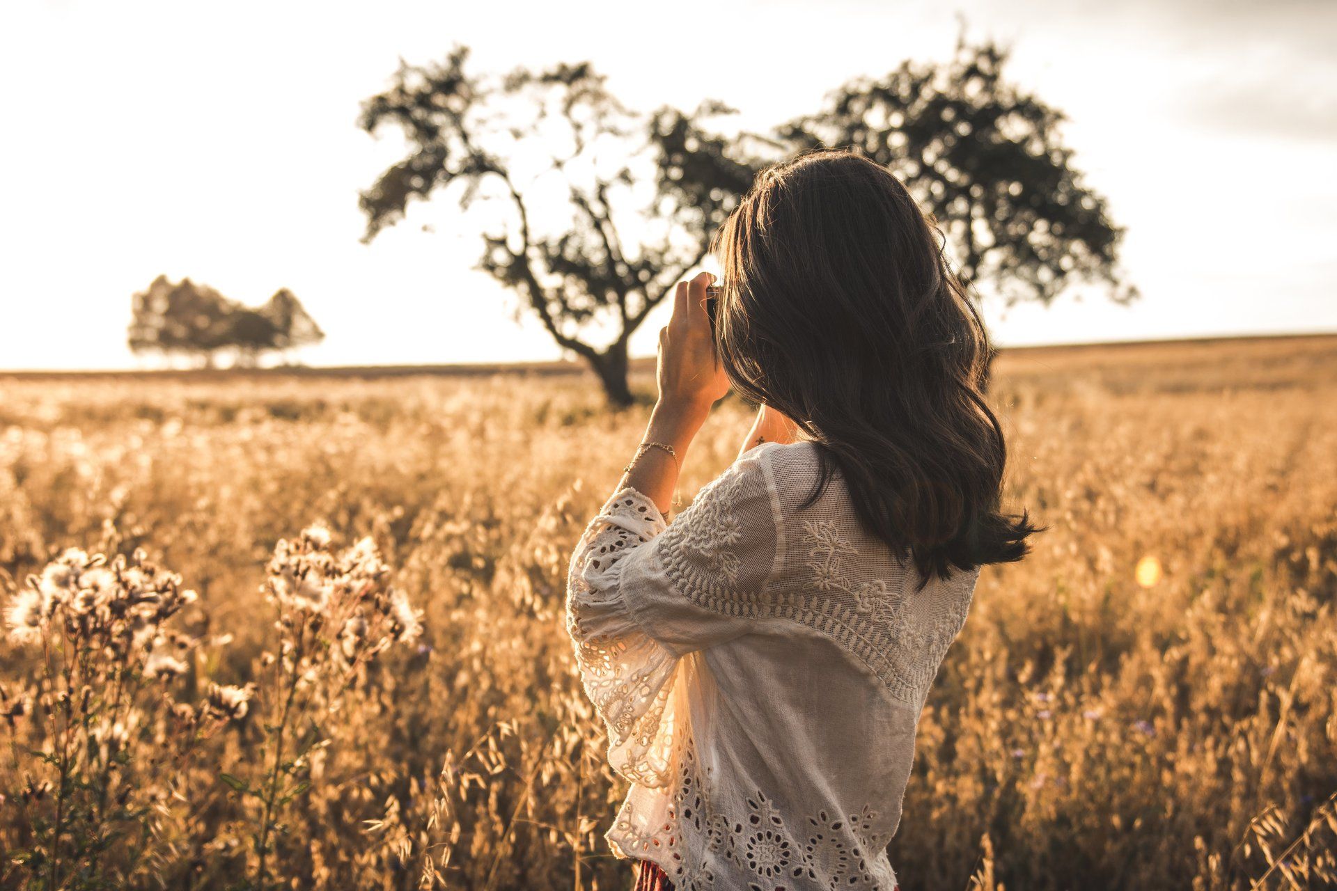 Frau in einem weißen Spitzentop macht ein Foto auf einem Feld mit goldenem Gras und Bäumen im Hintergrund.