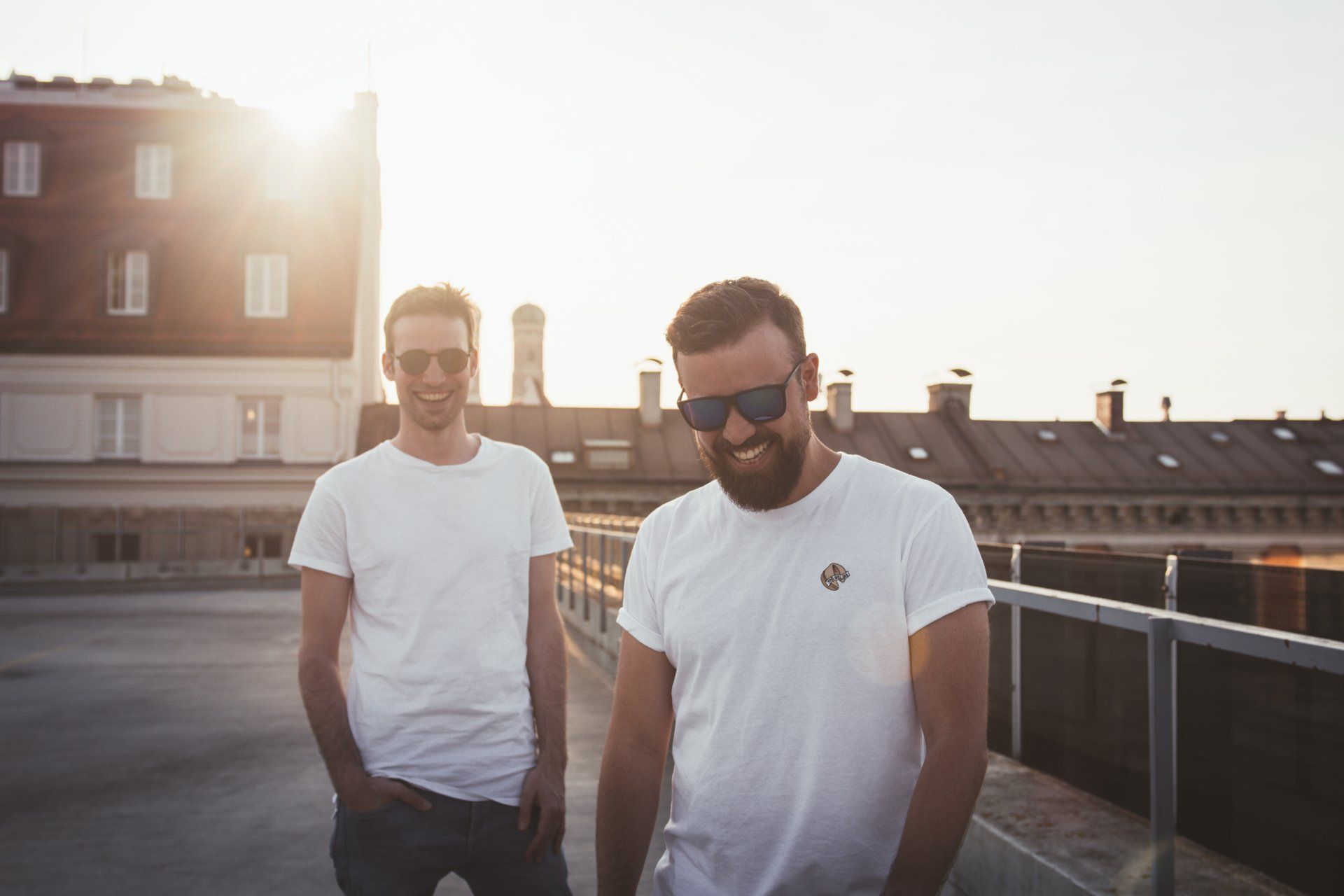 Zwei Männer mit Sonnenbrillen und weißen T-Shirts auf einem Dach, die im Sonnenlicht lächeln.