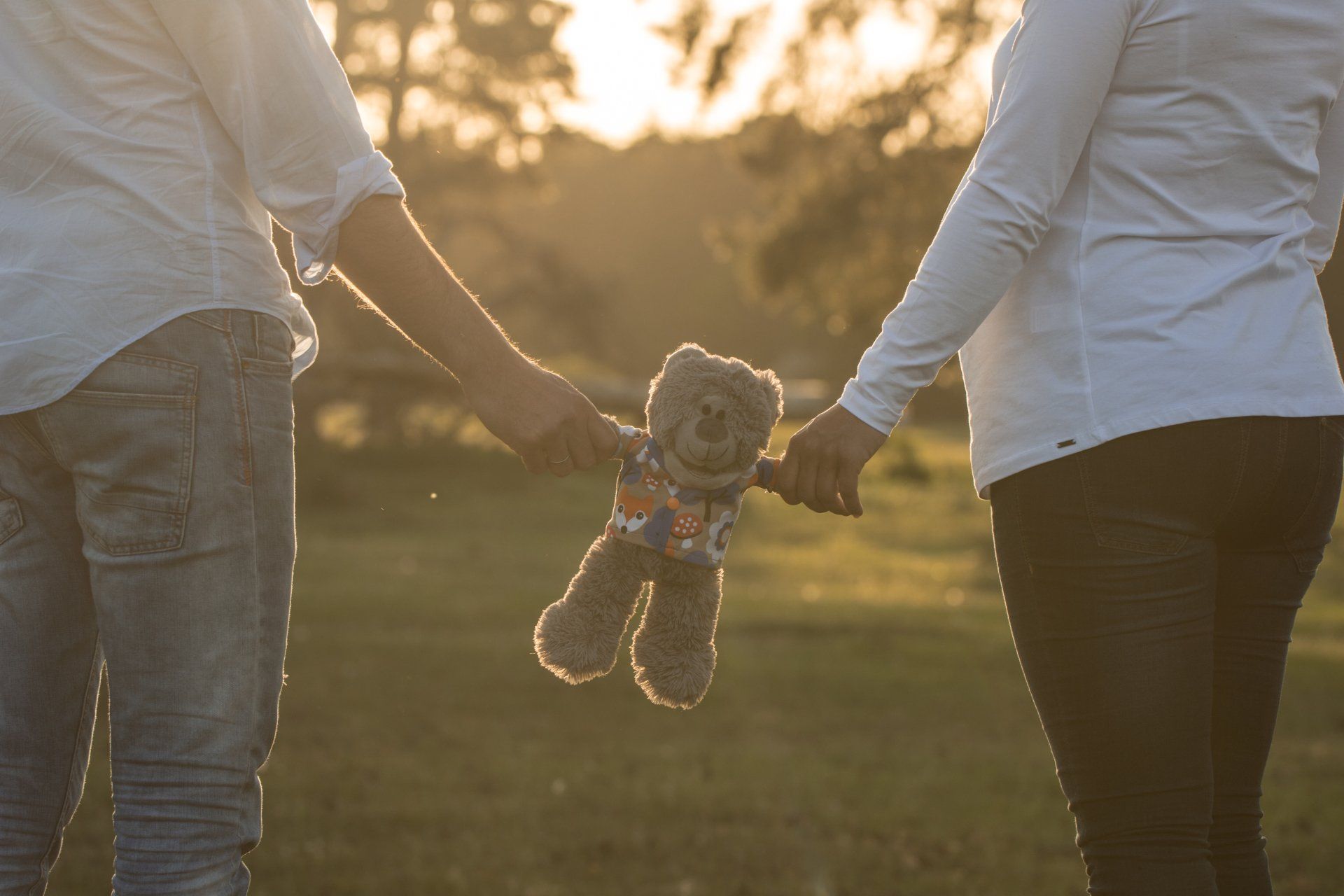 Paar hält Händchen, zwischen ihnen hängt ein Teddybär, im Gegenlicht des goldenen Sonnenlichts.