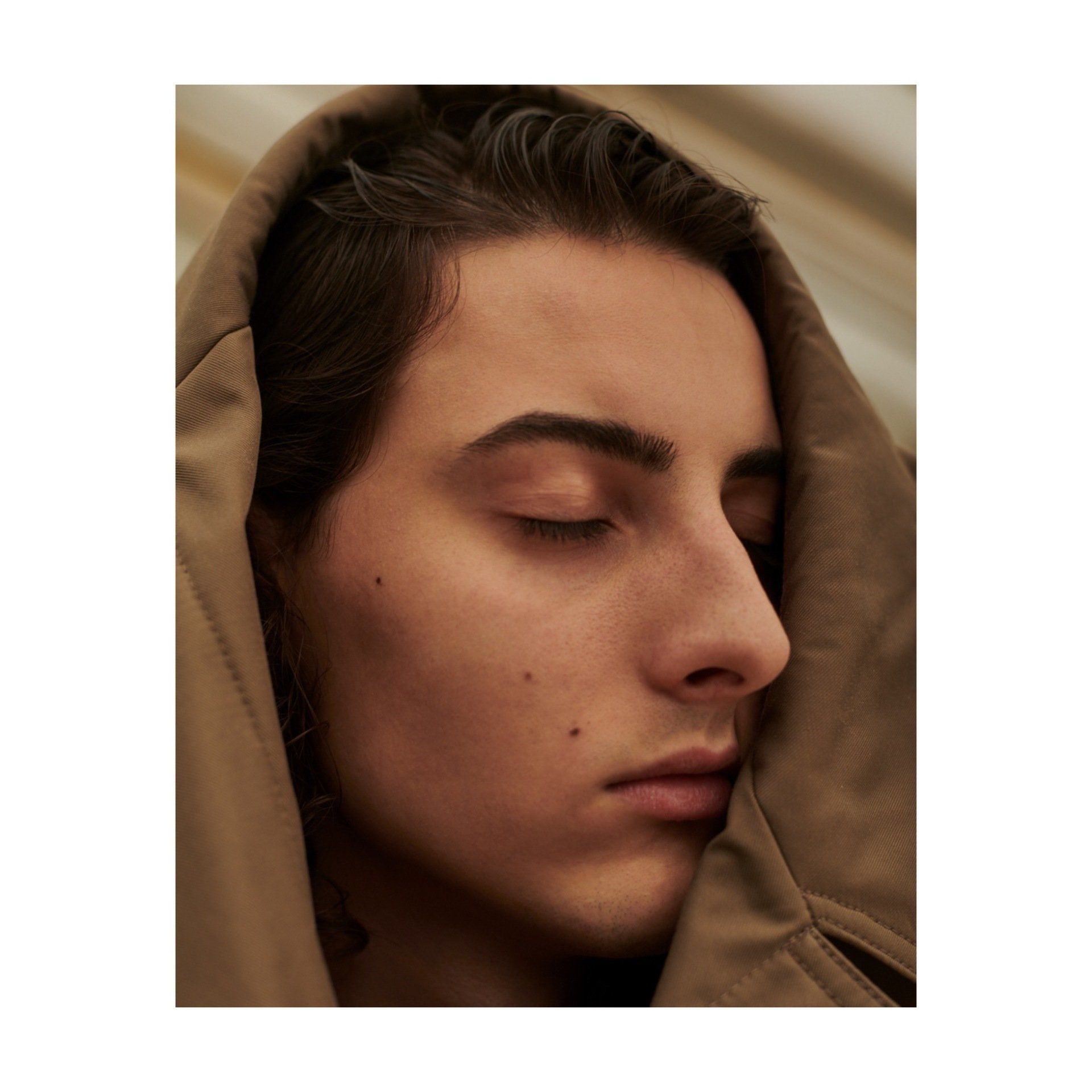 Person mit geschlossenen Augen, das Gesicht teilweise von einer braunen Kapuze bedeckt, Nahaufnahme.