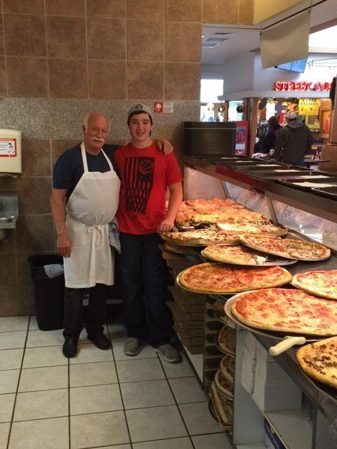 Two men are posing for a picture in front of a display of pizzas
