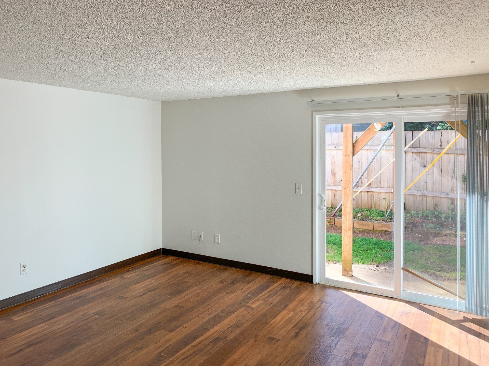An empty living room with hardwood floors and a sliding glass door leading to a backyard.