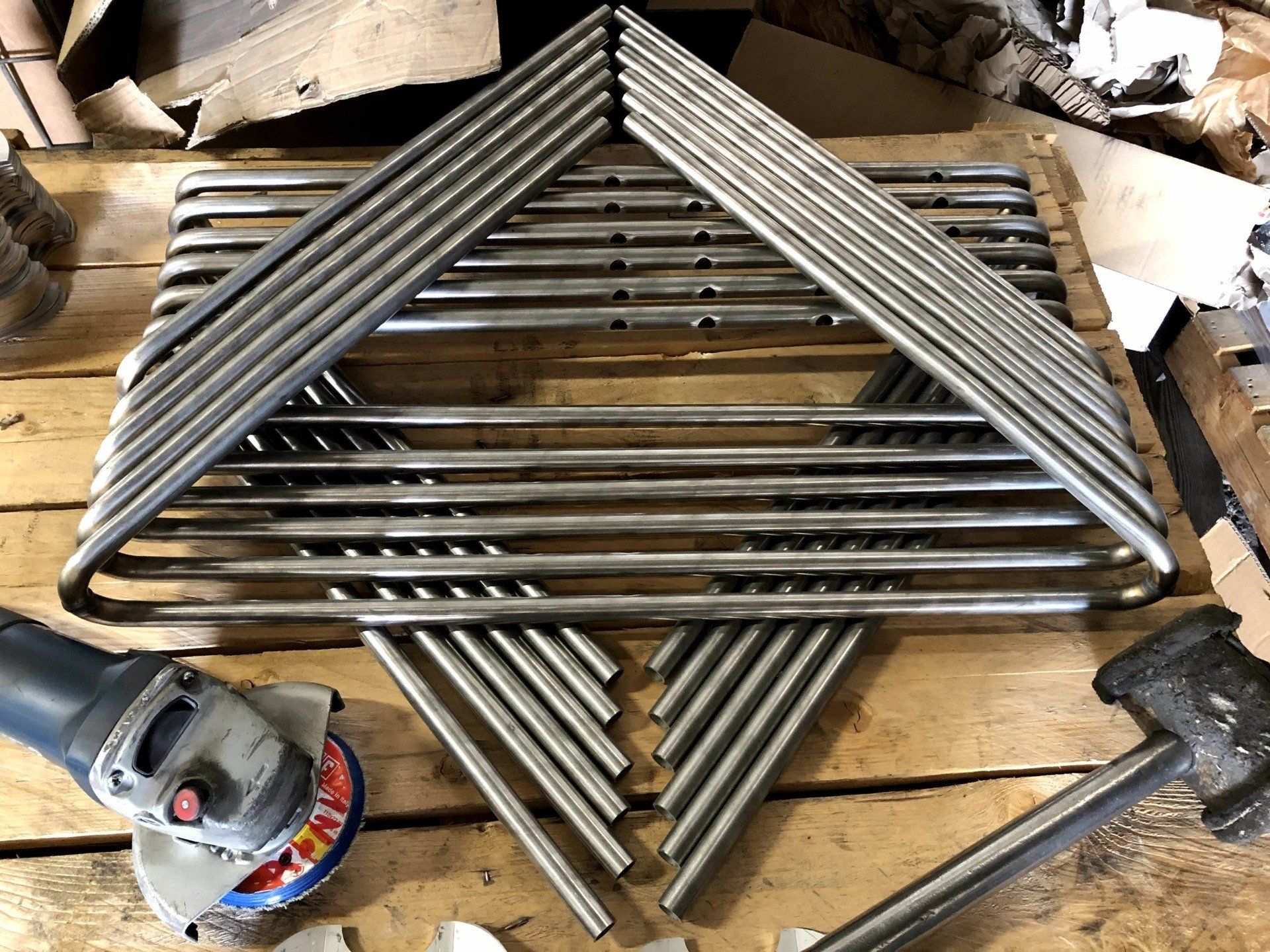 Tubi a U in acciaio inossidabile impilati e disposti secondo uno schema geometrico su un pallet di legno, con una smerigliatrice angolare e un martello.