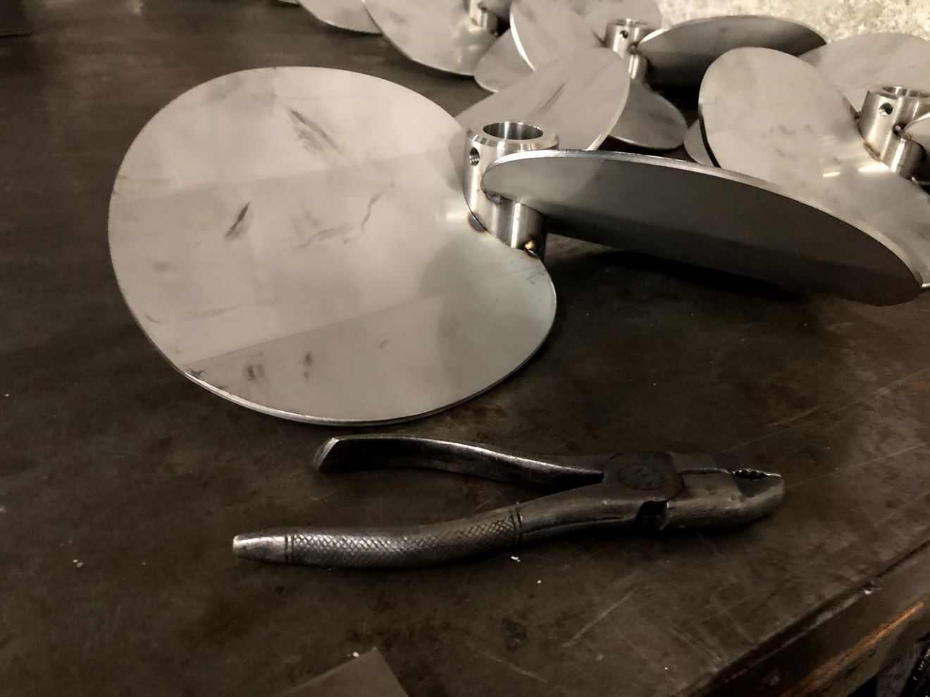 Diverse eliche metalliche a più pale sono appoggiate su una superficie di lavoro scura, accanto a un paio di pinze autobloccanti.