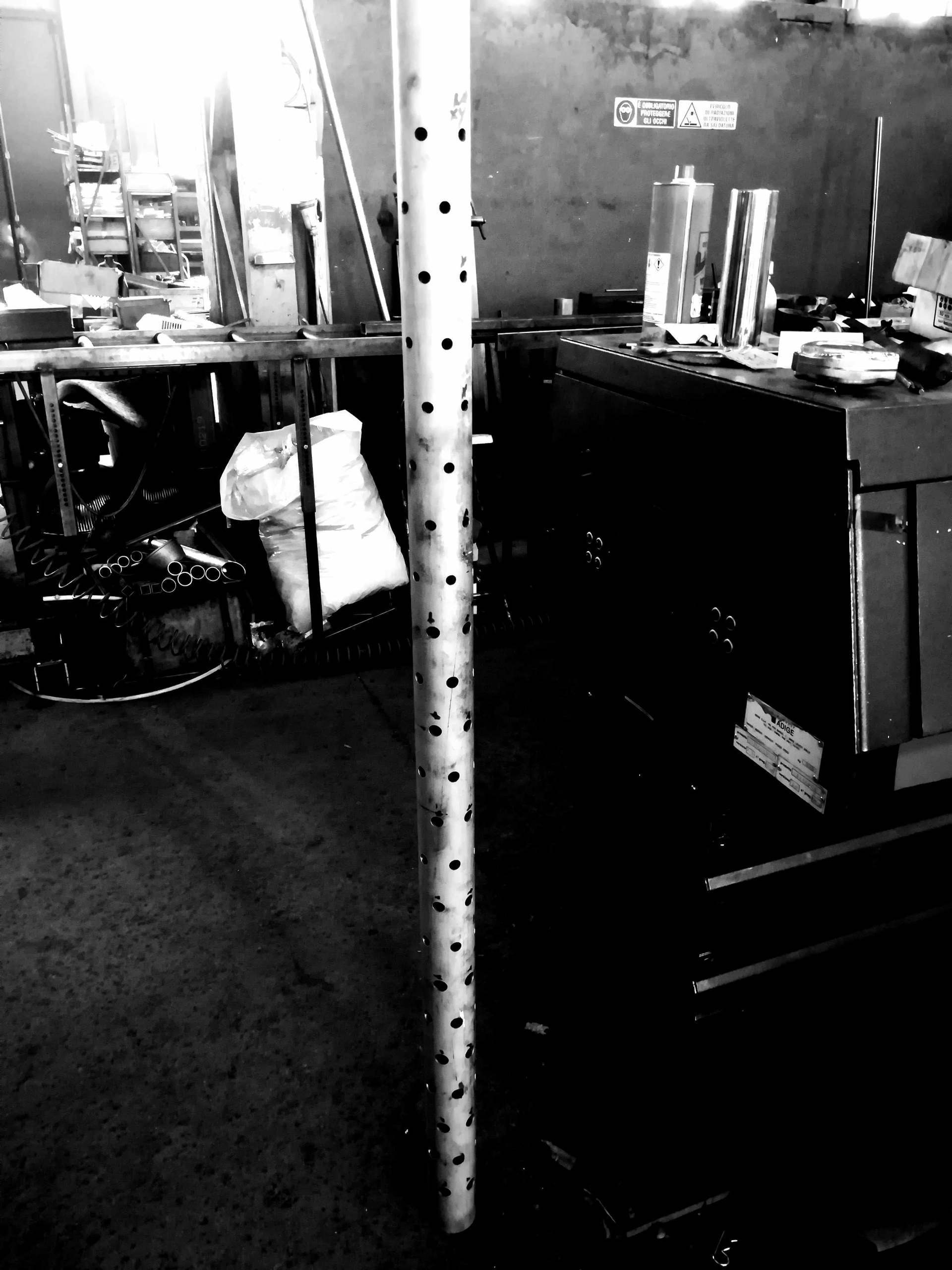 In un'officina industriale scarsamente illuminata e ingombro di attrezzi e componenti, si erge un palo metallico verticale con fori equidistanti.