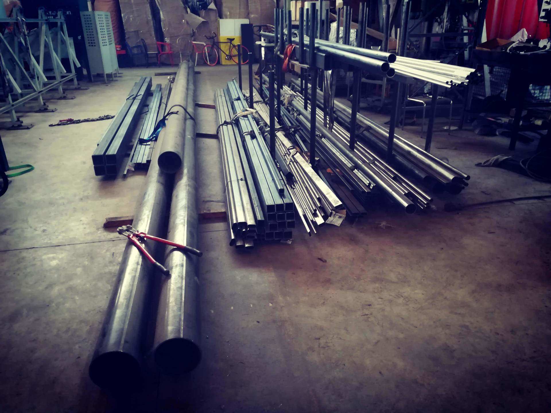 Pile di tubi metallici industriali e travi in ​​acciaio strutturale disposte sul pavimento in cemento di un'officina.