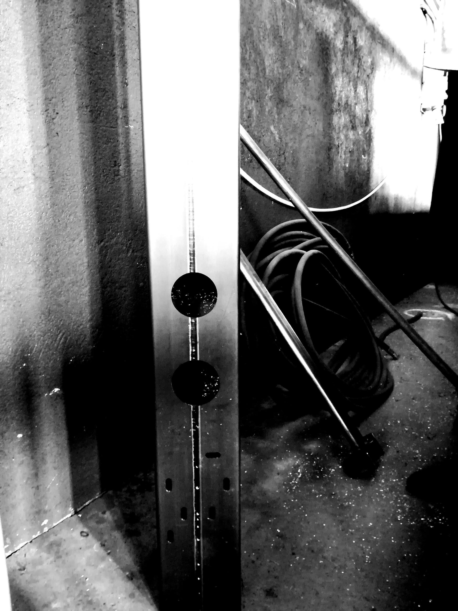 Un palo strutturale metallico verticale con due aperture circolari e diverse fessure, che si erge in uno spazio industriale scarsamente illuminato.