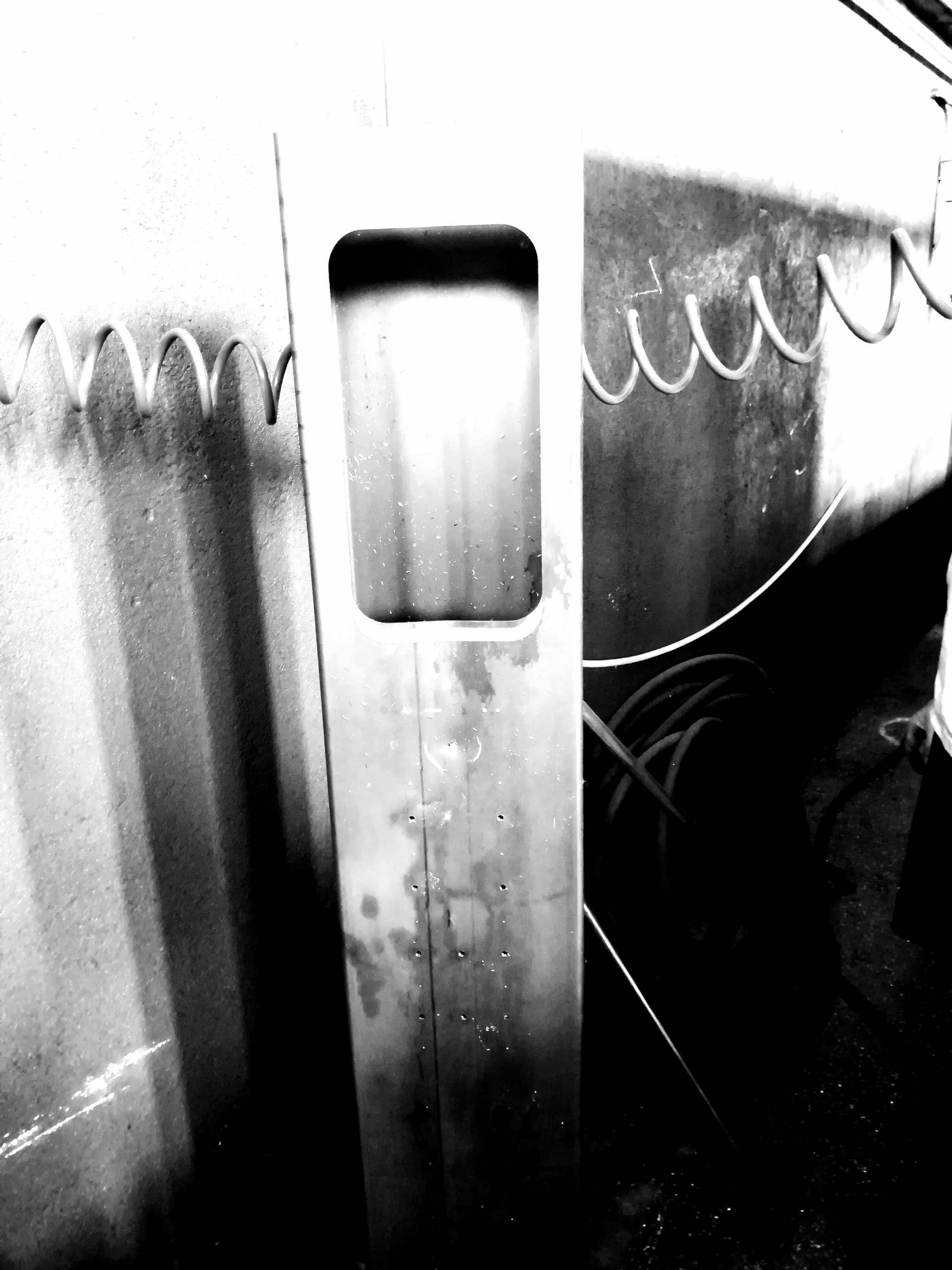 Un primo piano in bianco e nero ad alto contrasto di un palo metallico verticale con un'apertura rettangolare, posto davanti a una recinzione.
