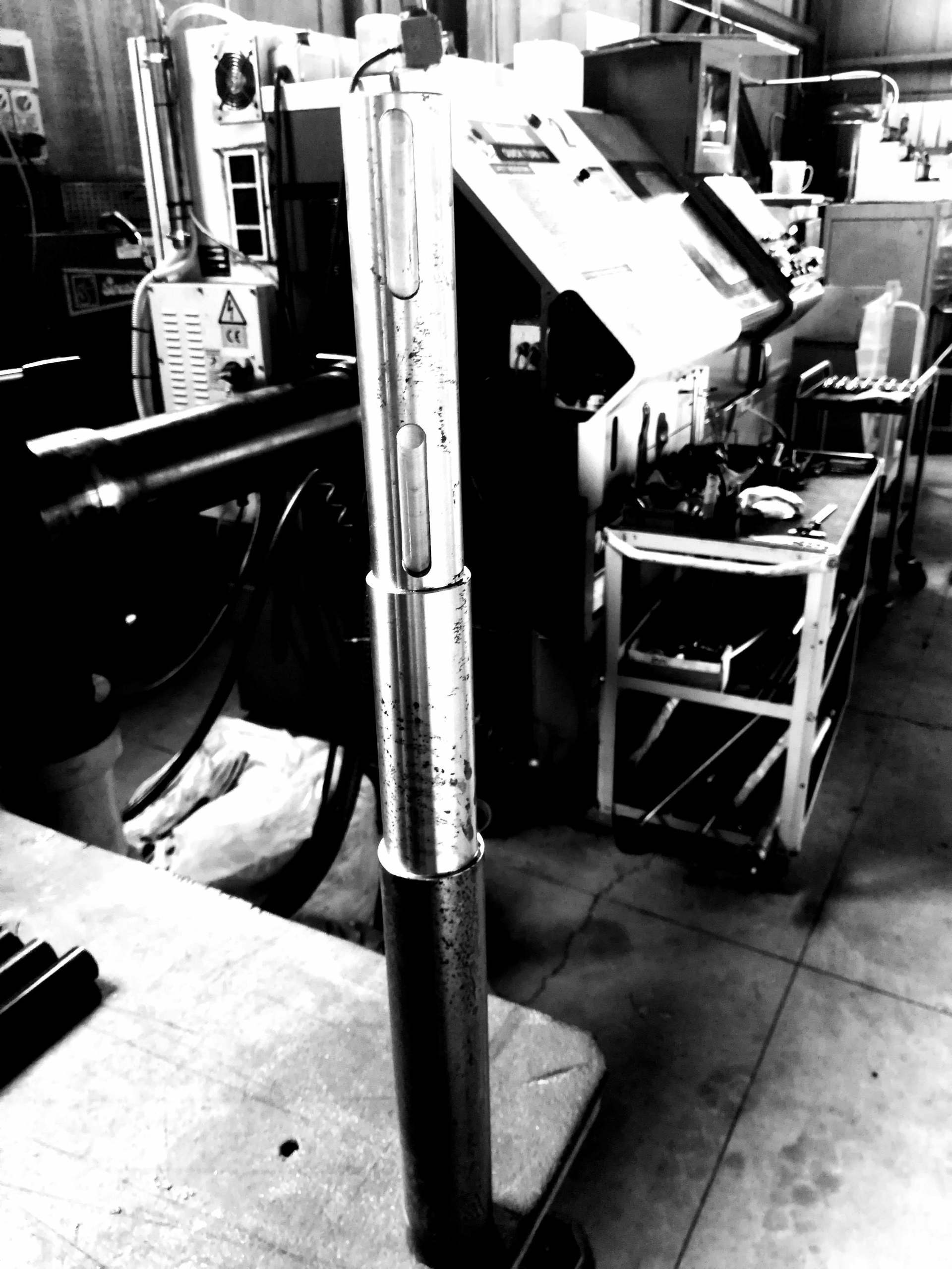 Un assemblaggio cilindrico metallico verticale in un'officina, sullo sfondo di macchinari e di un carrello metallico con ruote.