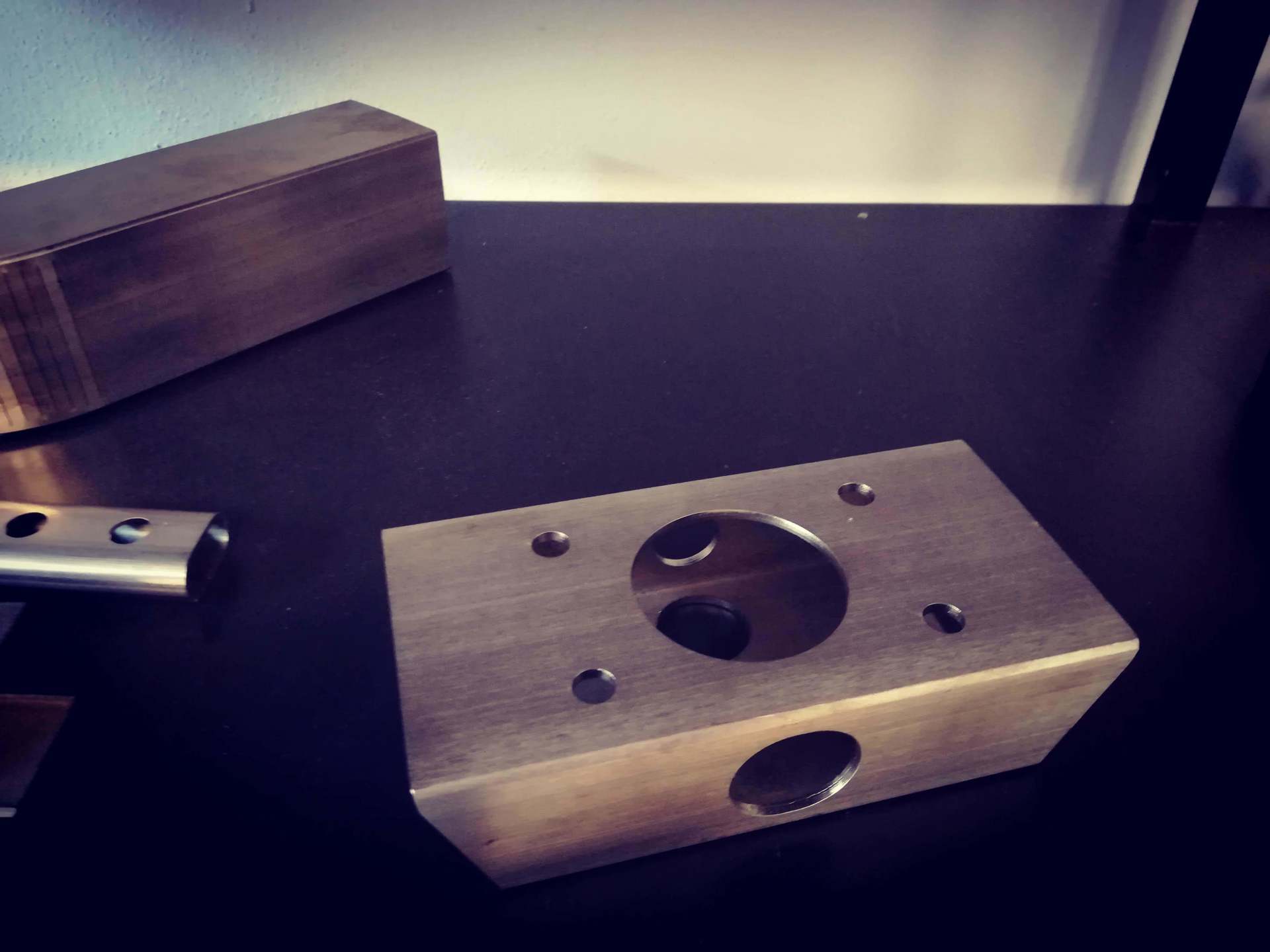Un blocco di metallo con un grande foro centrale e quattro fori di montaggio più piccoli è appoggiato su una scrivania scura accanto ad altri componenti metallici.