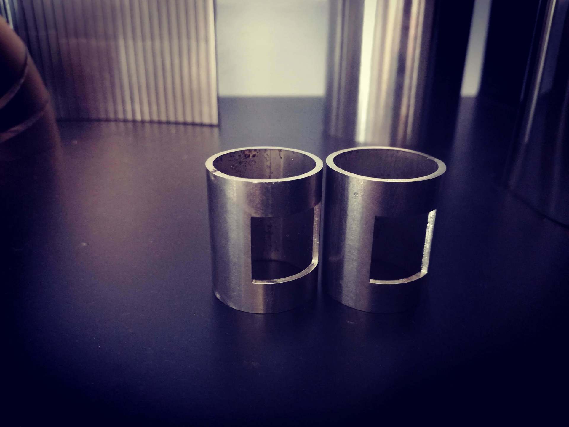 Due piccole guaine metalliche cilindriche con ritagli rettangolari, appoggiate su una superficie scura.