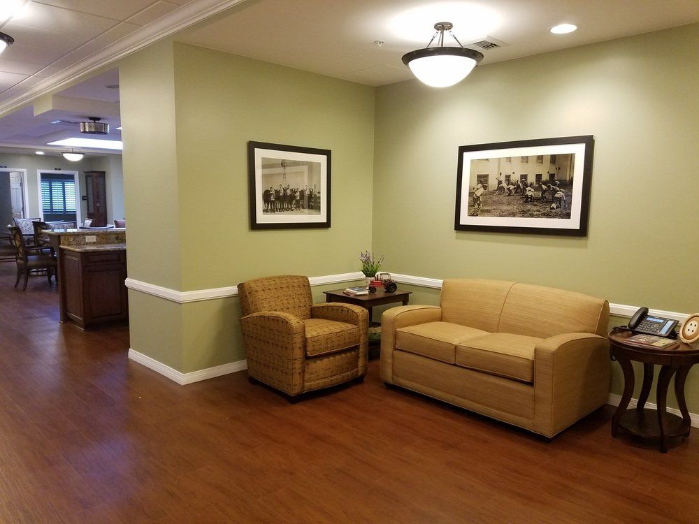 An indoor seating area: green walls, tan sofa and chair, framed art, and a table on wooden flooring.