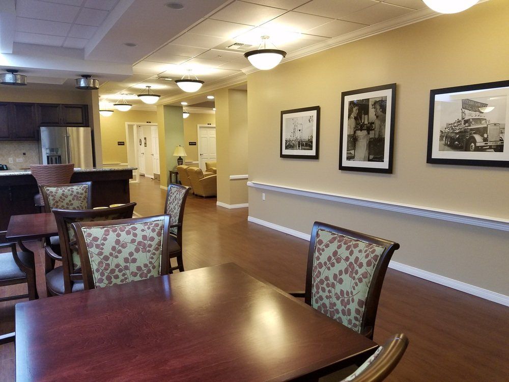 Interior of a long hall with seating, tables, and framed pictures; light tan walls and wood floors.