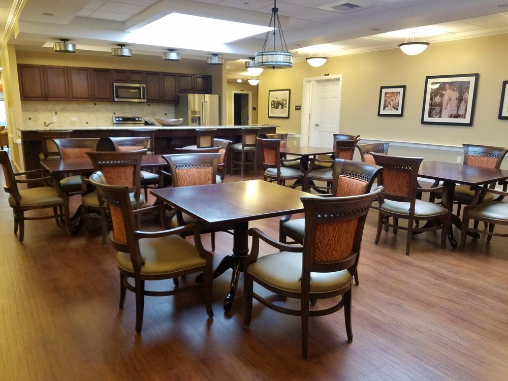 Interior of a dining area in a senior living facility; tables, chairs, and a small kitchen space.
