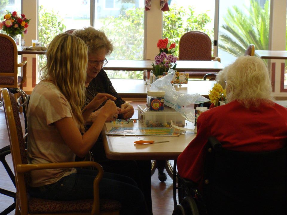 Three women crafting at a table in a sunny room. One woman is in a wheelchair.
