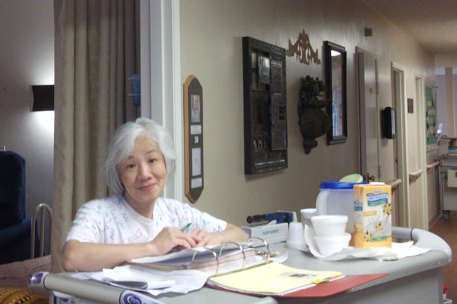 Woman with white hair at a desk smiles; beige walls, pictures, and food items nearby.