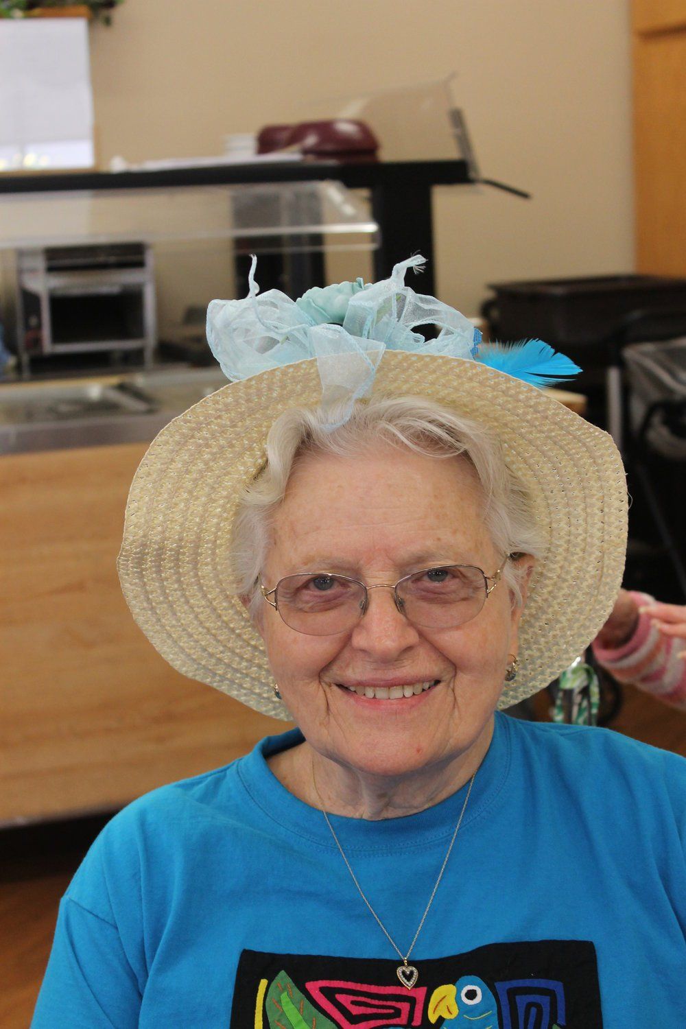 Elderly woman with glasses and straw hat, smiling, wearing a blue shirt, indoors.