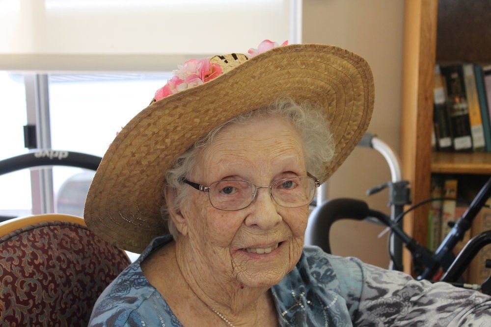 Elderly woman with glasses and straw hat smiling indoors.