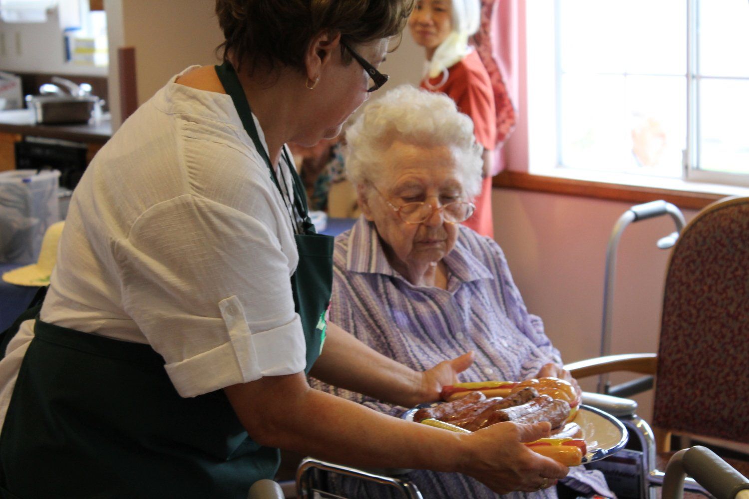 Woman in apron serves meal to elderly woman in wheelchair.