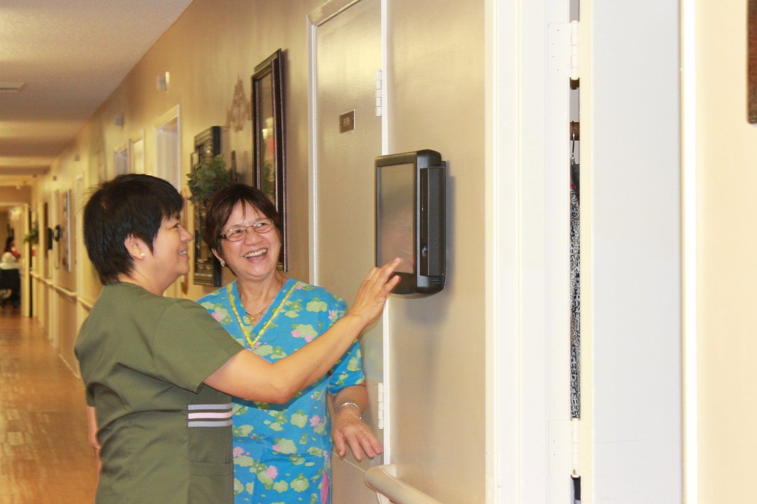 Nurse and patient laughing, interacting with a device on hallway wall.
