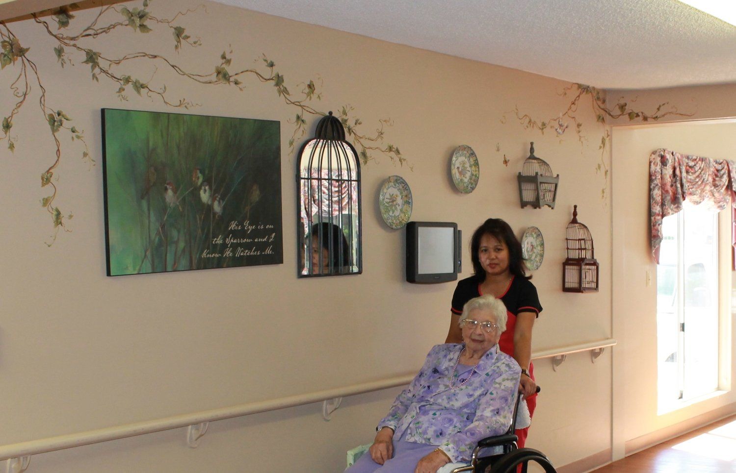Caregiver pushing elderly woman in wheelchair down decorated hallway.