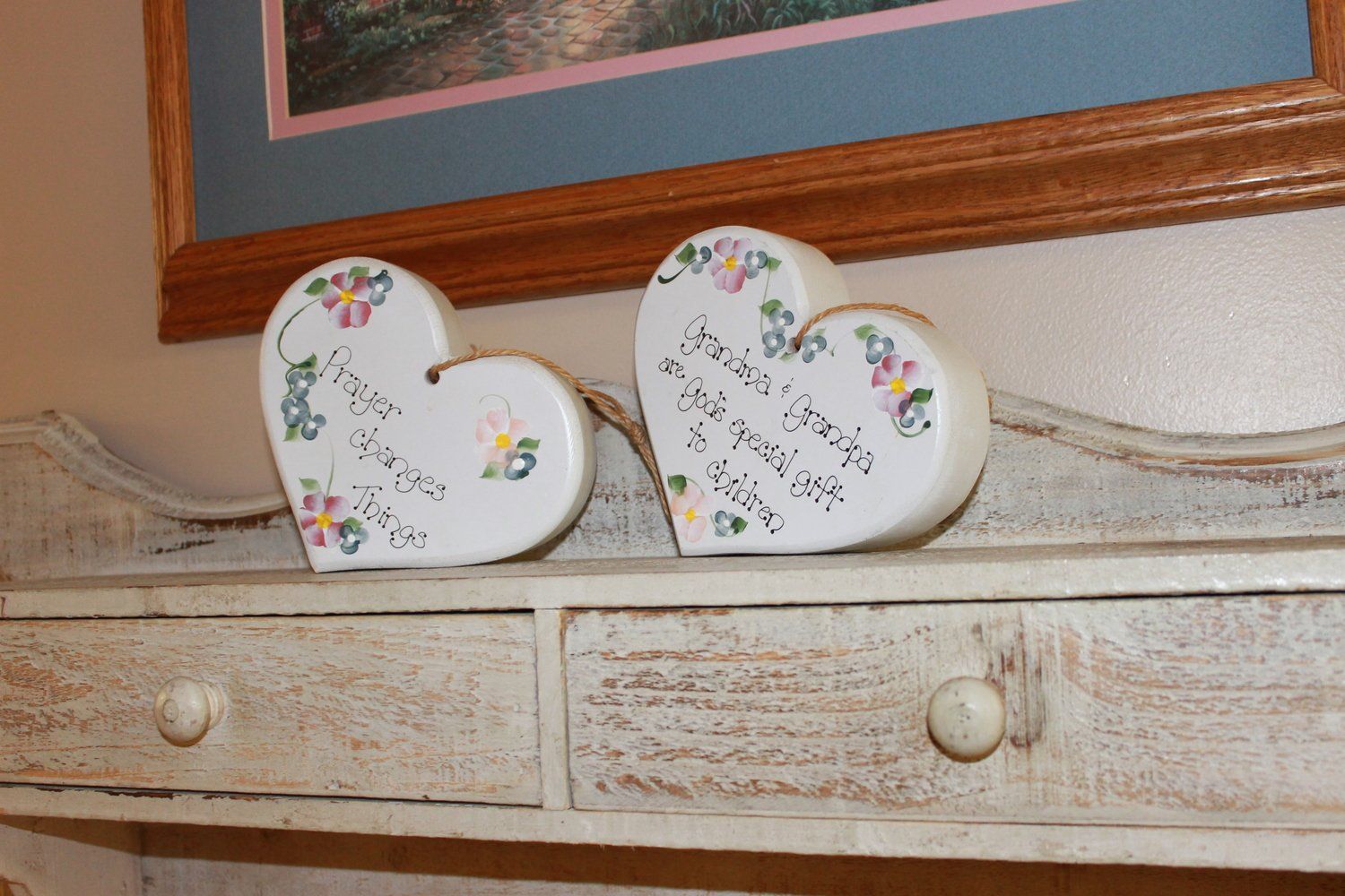 Two heart-shaped decorations with painted flowers and text on a white shelf, beneath a framed picture.