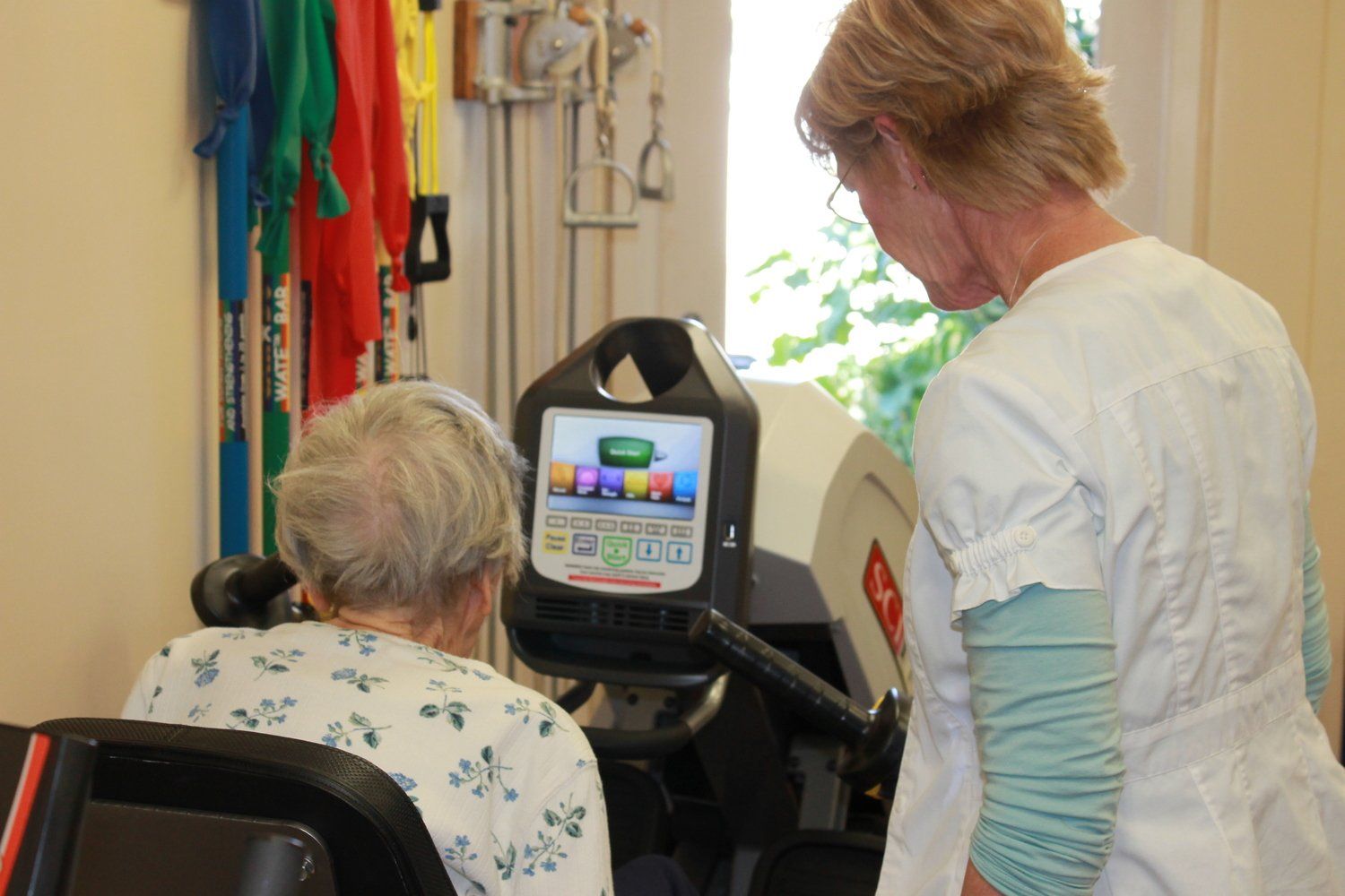 Elderly woman in wheelchair using exercise machine with assistance from a healthcare worker.