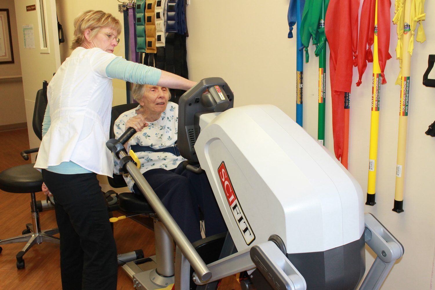 Therapist assists elderly woman using seated exercise machine in a rehabilitation room.
