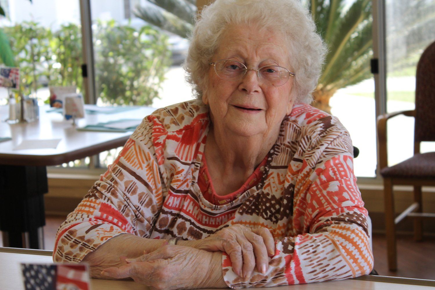 Elderly woman with white hair wearing glasses and a patterned shirt, seated at a table near a window.