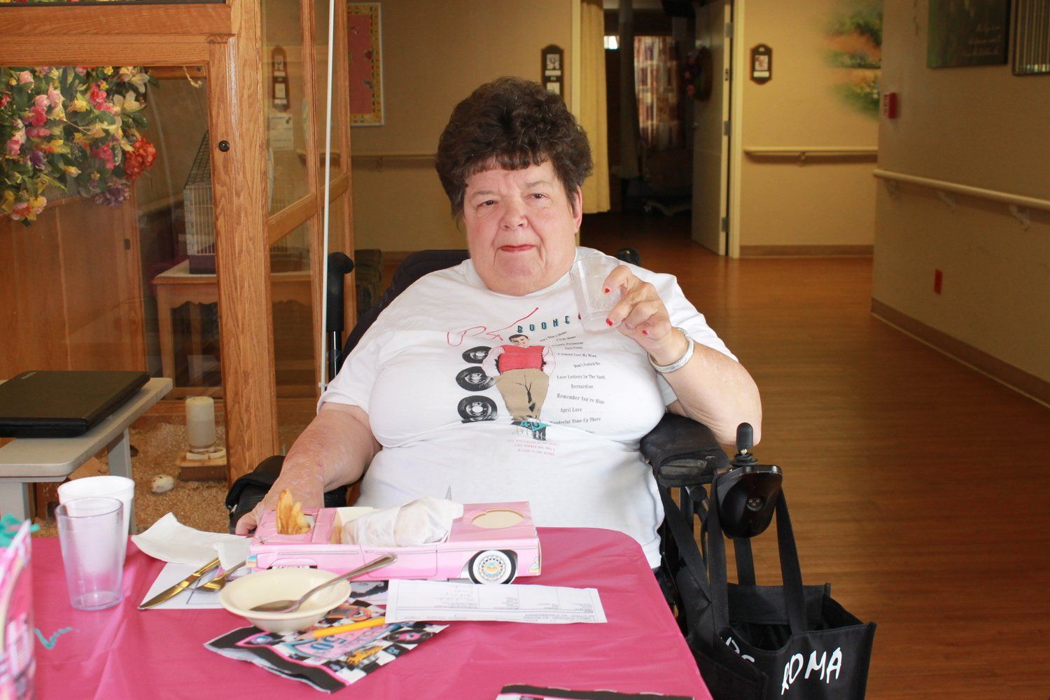 Woman in a wheelchair at a pink table with a milkshake, wearing a white t-shirt, inside a building.