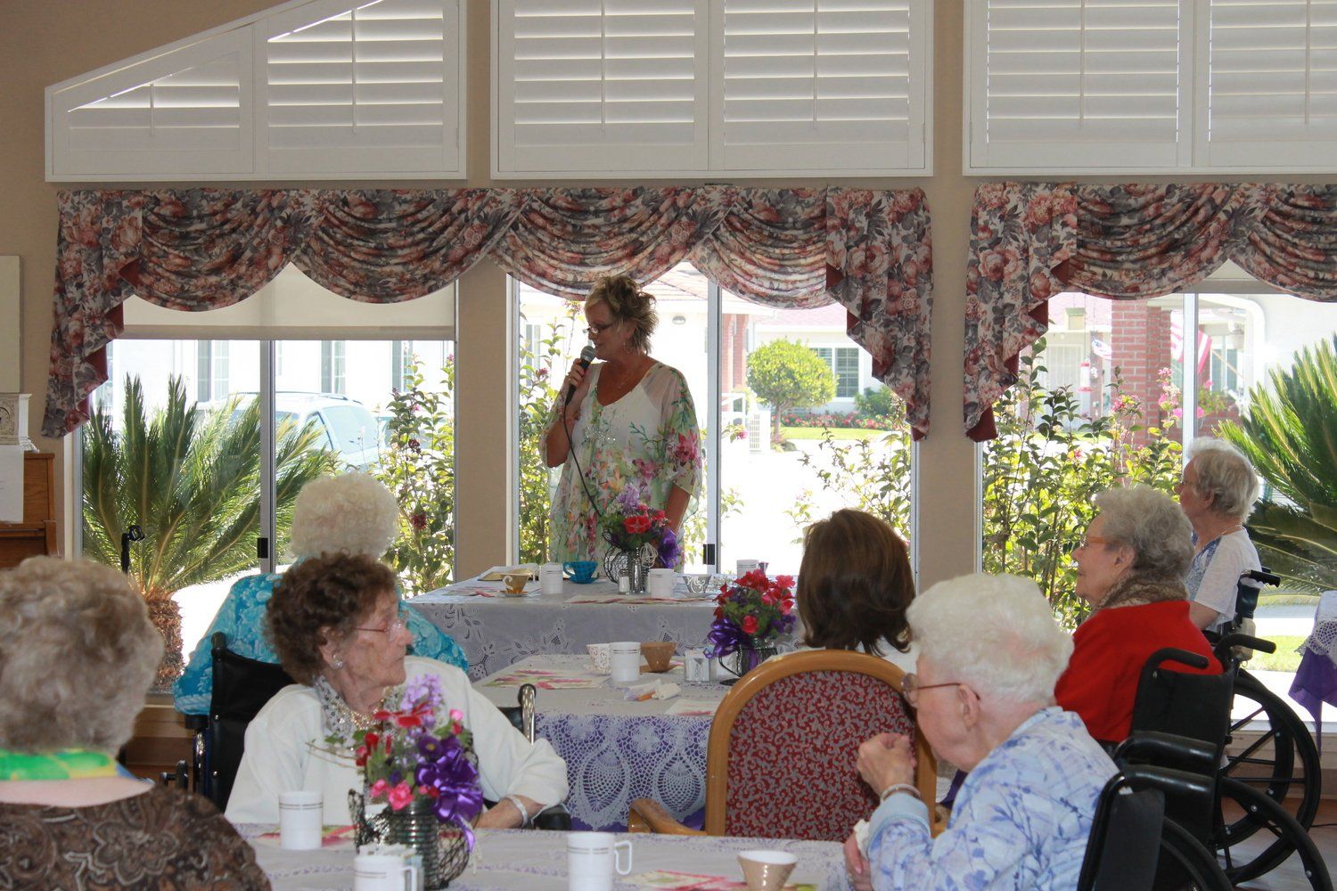 Woman speaks into a microphone at a table with older adults, in a bright room.