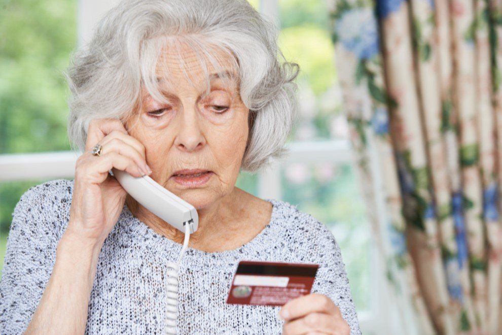 Elderly woman on phone looks distressed, holding a credit card near a window with floral curtains.