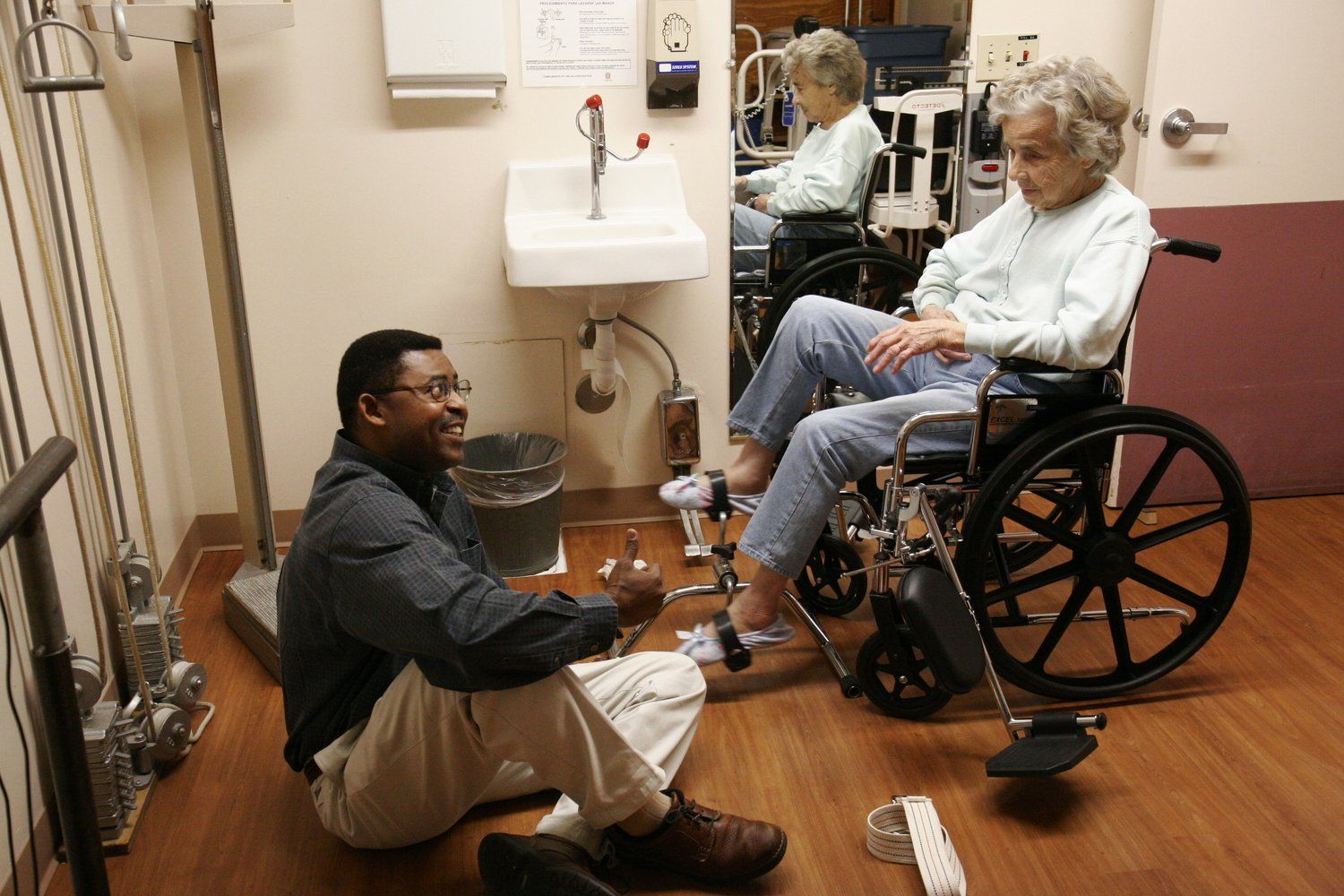 Man assisting elderly woman in wheelchair; rehab room. Man smiles while adjusting equipment near her feet.