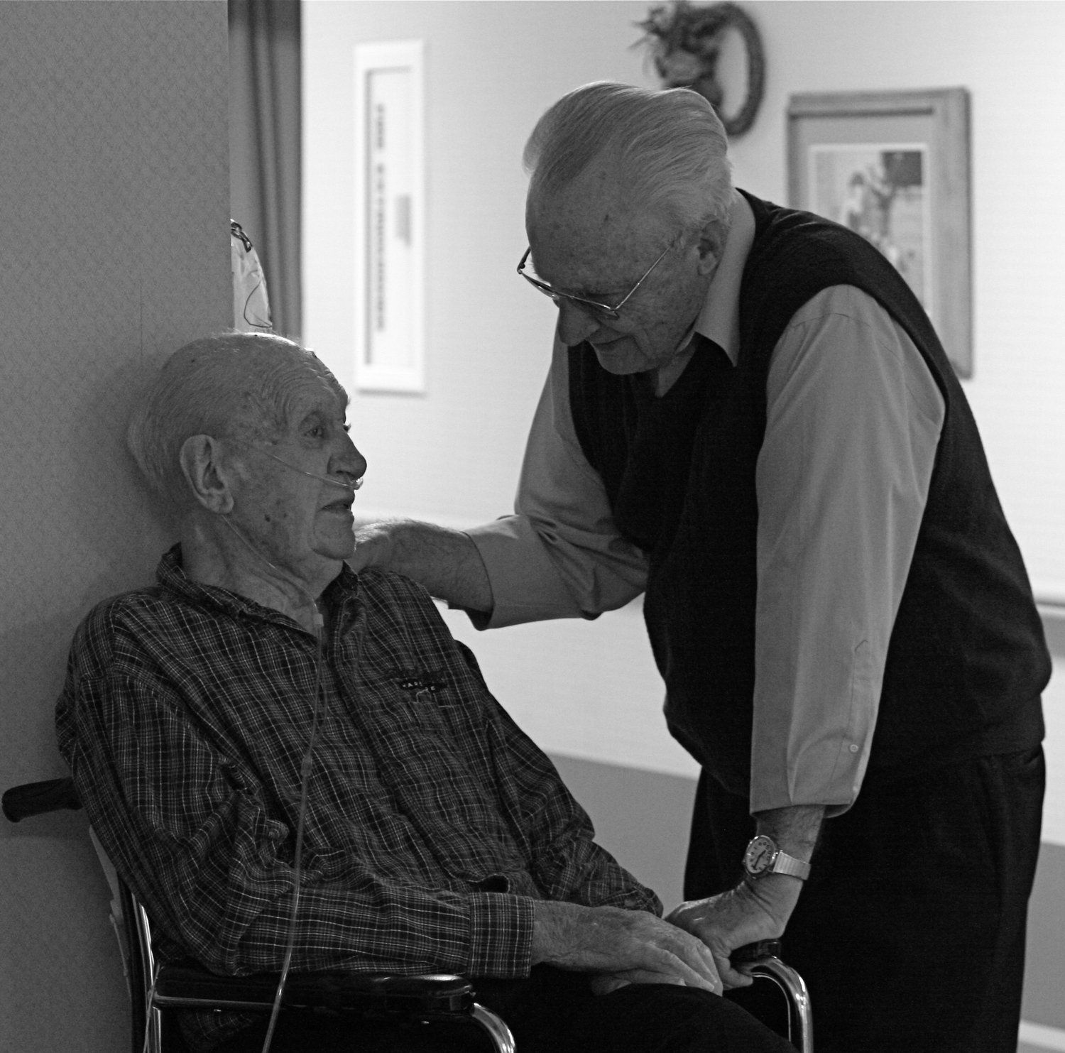Older man in wheelchair talking with another man who is standing and has his hand on the seated man's shoulder.
