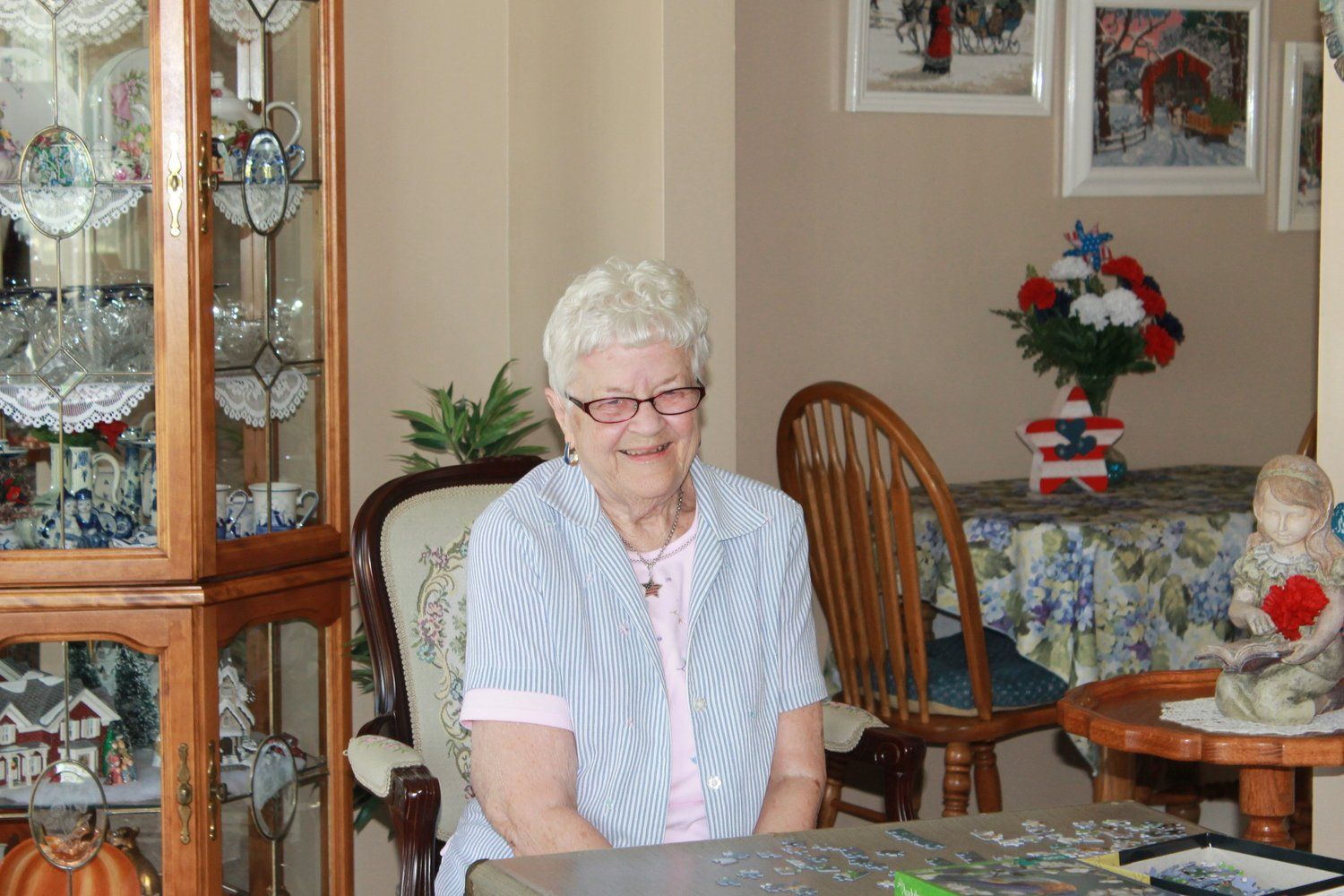 Elderly woman with glasses smiles, seated at a table, near a cabinet and decorated table with flowers.