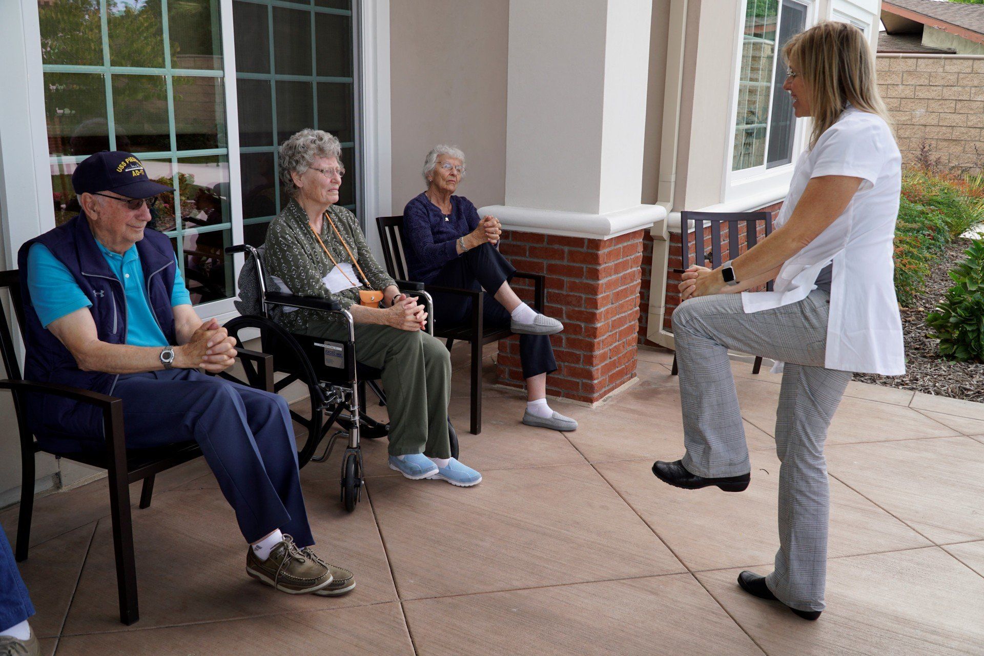 A woman demonstrates an exercise for three seated seniors on a patio.