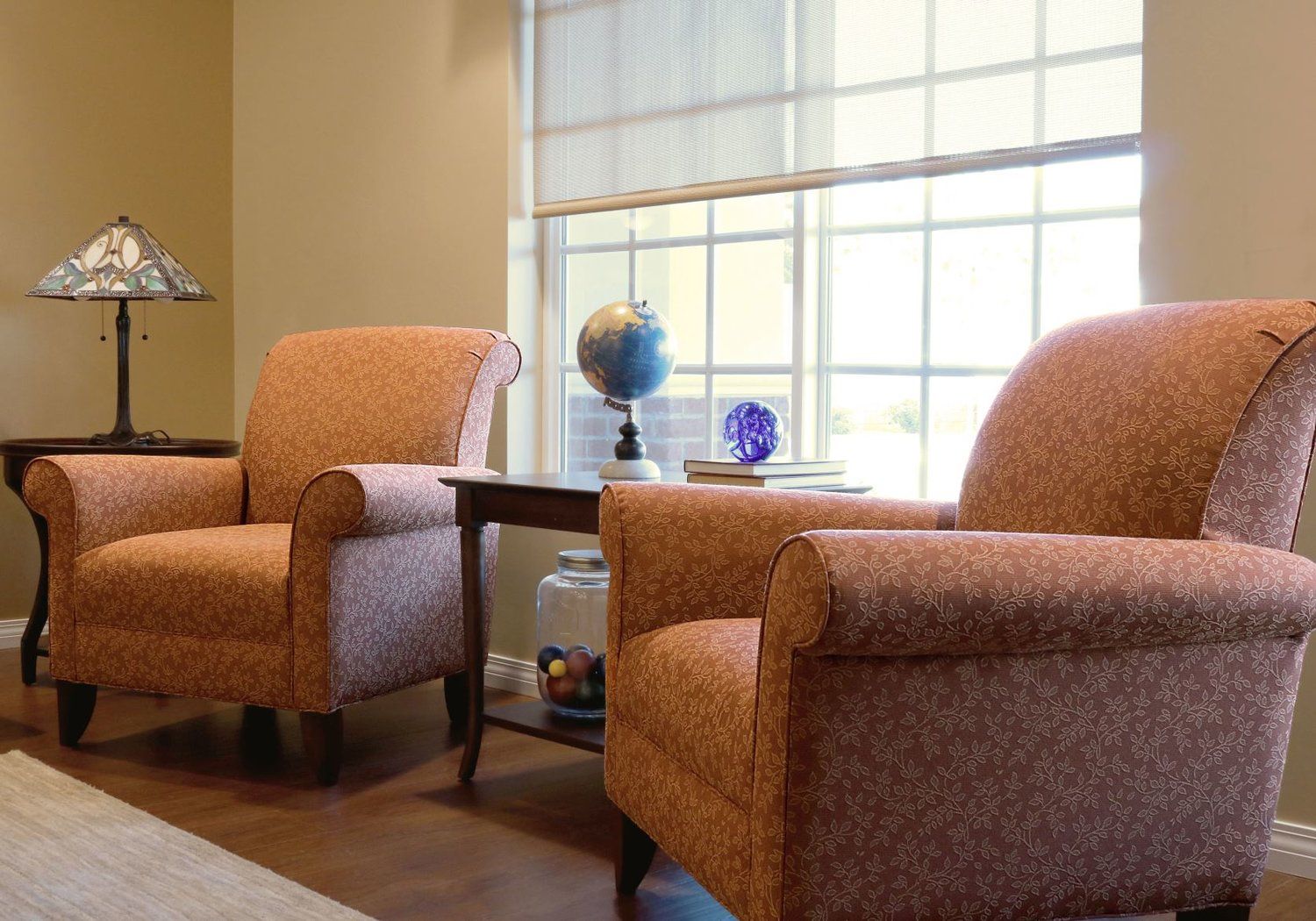 Two patterned armchairs face a window with a globe and decor on a table. A lamp sits on a side table.