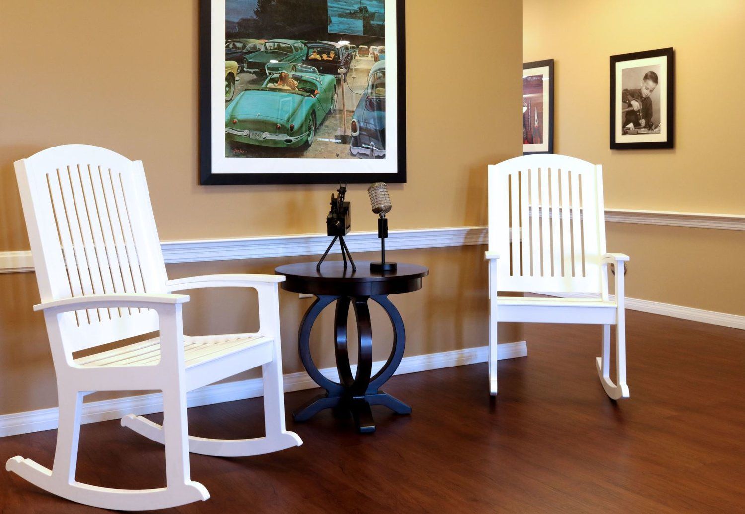 Two white rocking chairs beside a dark round table with microphones. Elvis portraits and a green car painting on wall.