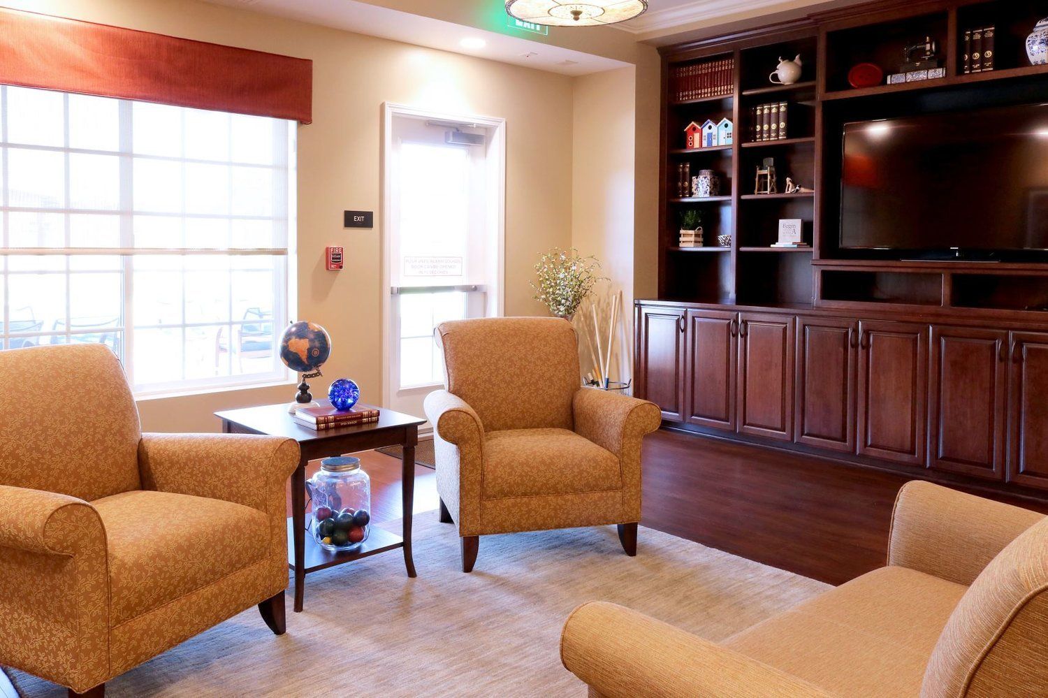 Cozy living room with two armchairs, a side table, and a built-in TV cabinet; sunlight streams through windows.