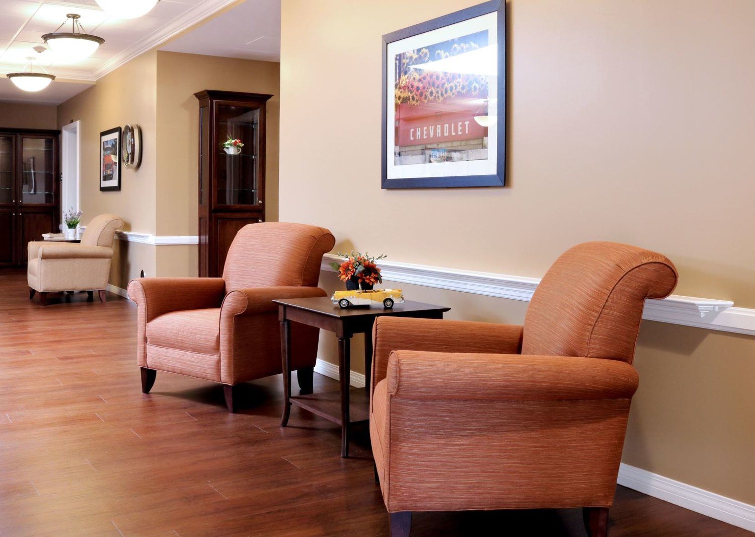 Two orange armchairs with a small table and a picture in a tan-walled hallway.